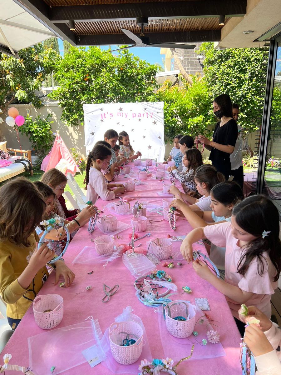 סדנת יצירה של לוכדי חלומות בהפעלת יום הולדת לבנות: בנות יוצרות סביב שולחן ורוד ארוך בחצר עם מדריכה