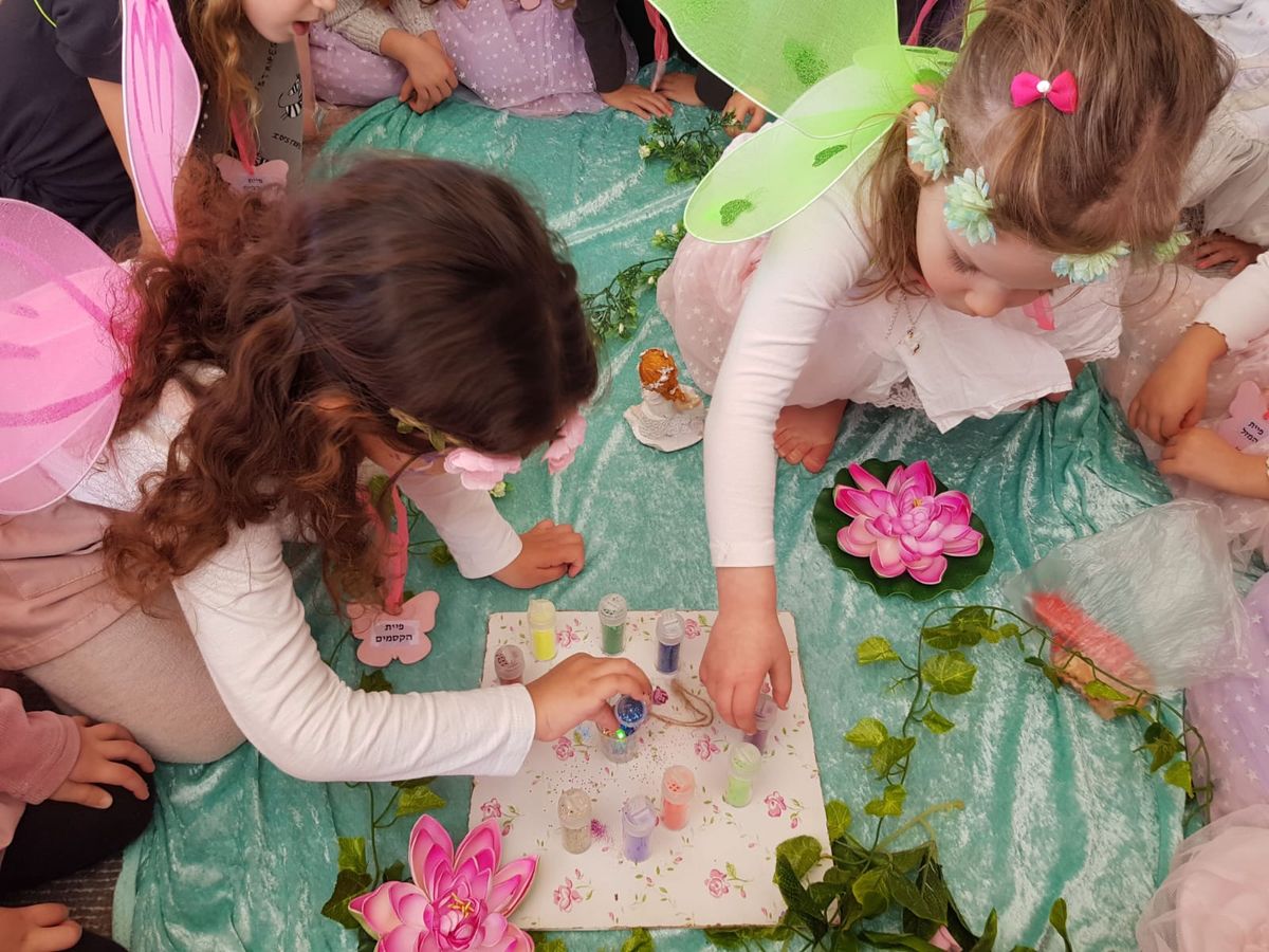בנות עם כנפי פיות רוכנות מעל בקבוקוני נצנצים צבעוניים על שטיח קסום בהפעלת יום הולדת לבנות