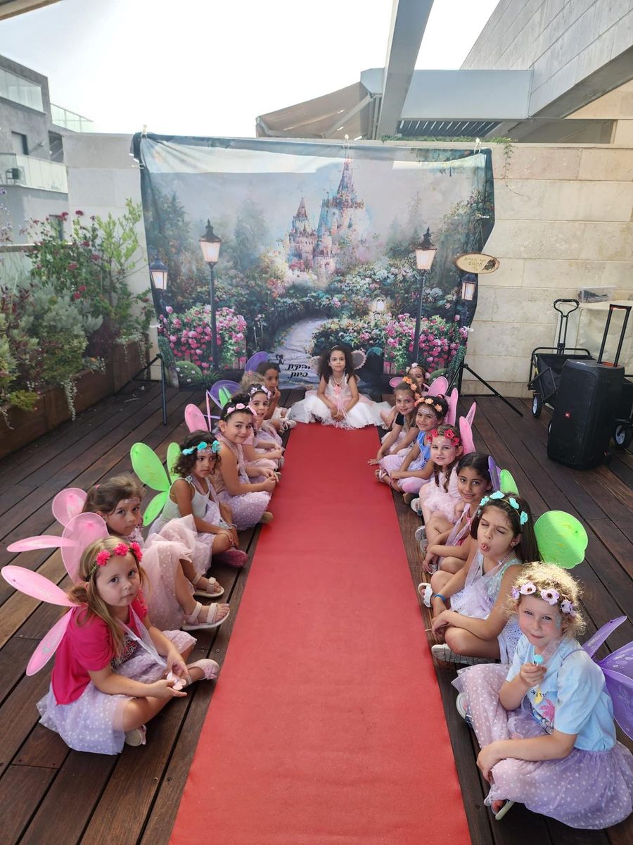 מסיבת נושא פיות ביום הולדת לבנות: בנות עם כנפיים וזרי פרחים יושבות לצד שטיח אדום מול תפאורת טירה קסומה