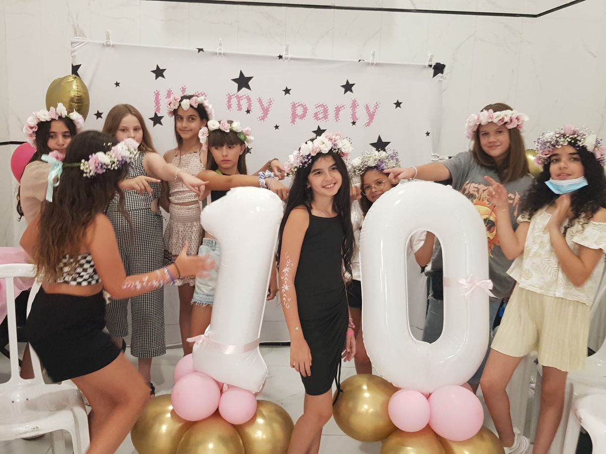 קבוצת ילדות עם זרי פרחים חוגגות יום הולדת בסלון מקושט, עומדות מסביב לבלוני מספר גדולים על רקע קיר עם כיתוב חגיגי וכוכבים