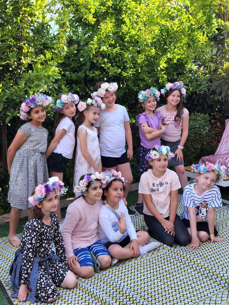 קבוצת ילדות בגינה חובשות זרי פרחים צבעוניים שיצרו בעצמן בסדנת שזירת פרחים, מחייכות יחד על שטיח בחצר מוקפת ירק