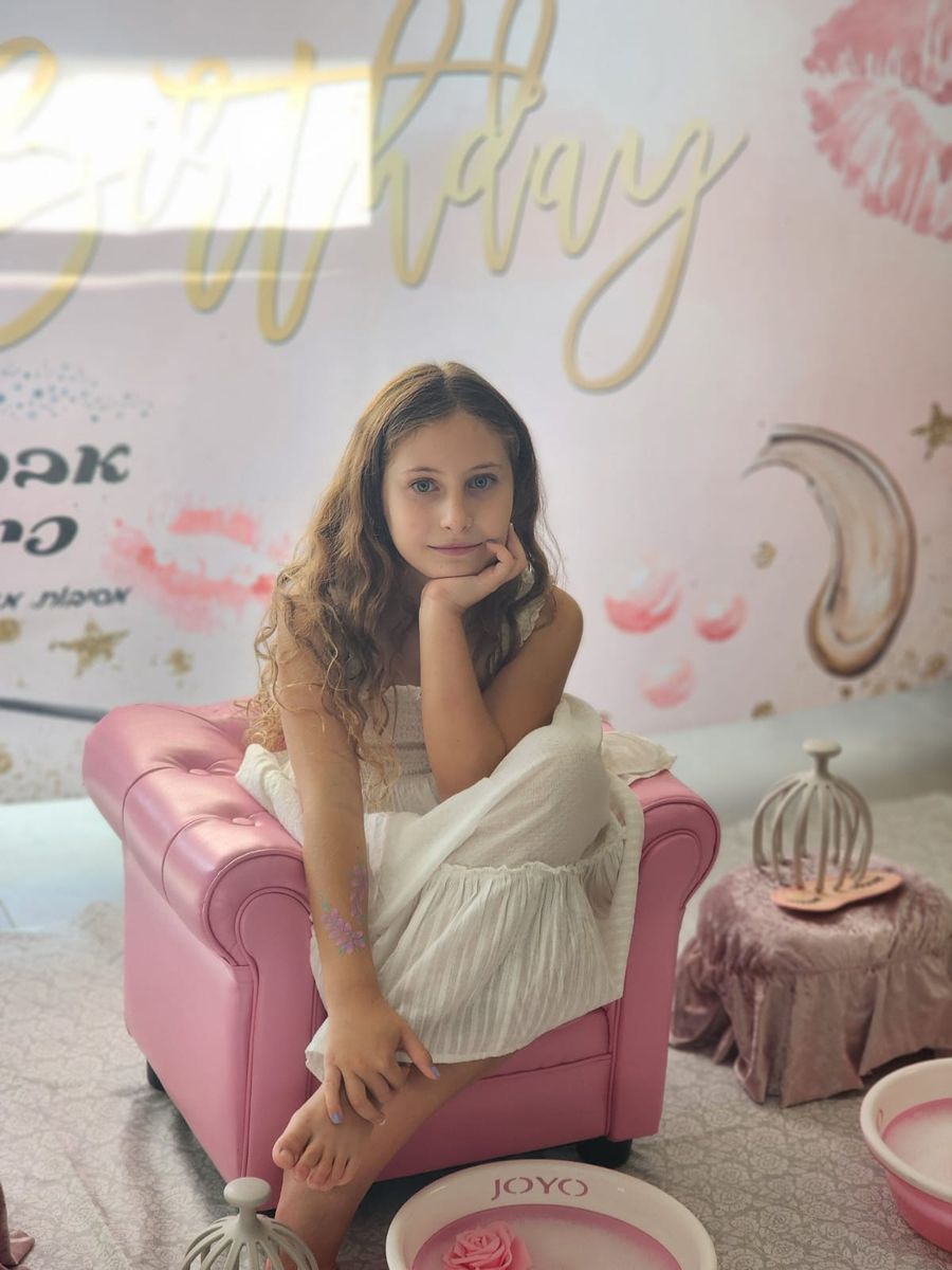 ילדה יושבת בנוחות בכורסה רוזה בקונספט יום הולדת ספא, עם קערת טבילת רגליים וכובע מסאז' לצידה
